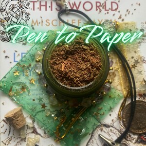 May include: A small green glass jar filled with a brown herbal blend, positioned on a green organza bag with gold stars. The scene includes a black cord necklace with a silver bird skull pendant, a cork stopper, and a metallic coin on paper.