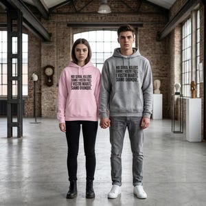 Può includere: Una felpa con cappuccio rosa chiaro e una felpa con cappuccio grigia, entrambe con tasca a marsupio e il testo nero: NOI SERIAL KILLERS SIAMO I VOSTRI FIGLI, I VOSTRI MARITI, SIAMO OVUNQUE.