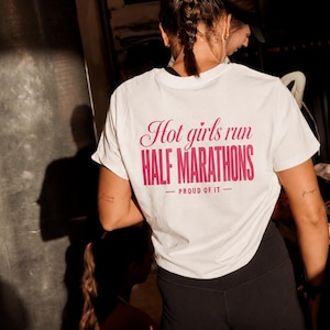 Peut inclure: Un t-shirt blanc à manches courtes avec un texte rose au dos indiquant Hot girls run HALF MARATHONS — PROUD OF IT — associé à un short de sport noir. Le t-shirt présente une encolure ronde et une coupe standard, porté avec un short noir.