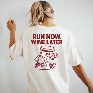 May include: Cream-colored t-shirt with the text "RUN NOW, WINE LATER" in burgundy. A cartoon wine glass character is running below the text. The shirt is a casual, oversized style.