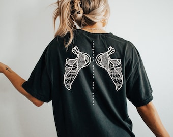 Può includere: Maglietta nera con una grafica bianca di due scarpe da arrampicata che formano ali. Il testo "climbing gives you wings" è stampato verticalmente sul retro della maglietta.