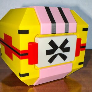 May include: A geometric, multi-colored box with a yellow body, pink top, and a white panel. The yellow sections have red and black stripes. The white panel features a black symbol. The box is on a wooden surface.