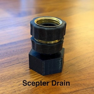 Op de afbeelding: Een zwarte Scepter Drain fitting met goudkleurige metalen accenten. De fitting heeft een zeshoekige basis en een schroefdraad bovenkant. De tekst "Scepter Drain" is in het wit op het oppervlak gedrukt.