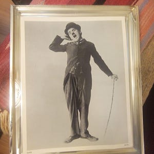 May include: Framed black and white photograph of Charlie Chaplin in character, wearing a bowler hat, suit, and holding a cane. The image captures Chaplin yawning with one arm raised, a classic pose.