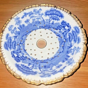 May include: A white porcelain bowl with a blue and gold design. The bowl has a perforated center and a scalloped rim.
