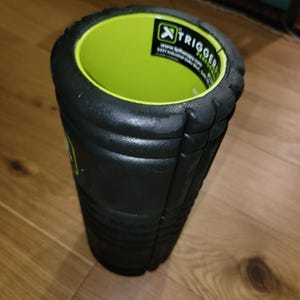 May include: Black foam roller with a bright green interior. The roller has a textured surface and the text "X TRIGGER PERFORMANCE" is visible on a white label. This fitness tool is designed for muscle recovery and self-massage.
