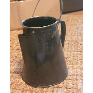 May include: A black enamelware coffee pot with a spout and handle. The pot has a speckled pattern and a silver rim. The handle is attached to the side of the pot. The pot is sitting on a woven surface.