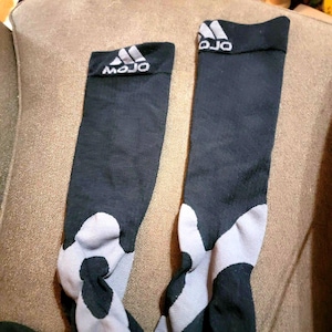 May include: A pair of black and grey athletic socks. The socks have a white and grey pattern on the foot and a black cuff with the word "MOJO" and a logo in white.