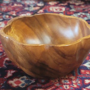 Puede incluir: Un cuenco de madera con borde ondulado. El cuenco es de madera marrón claro con un patrón de vetas visible. El cuenco está vacío y parece nuevo. Probablemente se utiliza para servir alimentos o como objeto decorativo, con un diámetro de unos 25 cm.
