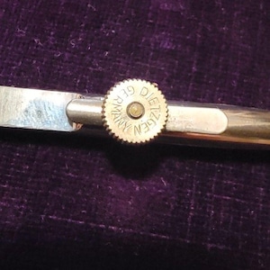 May include: A silver-toned metal tie clip with a circular, ridged center piece. The center piece has the words "DIETZGEN GERMANY" engraved around the edge. The clip is on a dark purple velvet surface.