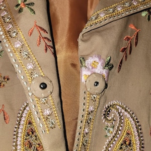 May include: Close-up of a tan jacket with intricate floral and paisley embroidery in shades of pink, green, and gold. The jacket features two round buttons and detailed stitching along the edges, showcasing a bohemian style.