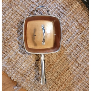 Może przedstawiać: Kwadratowa patelnia Copper Chef z miedzianym wnętrzem oraz srebrnym uchwytem i pokrywką. Patelnia stoi na brązowym, tkanym dywanie. Patelnia ma długi uchwyt i mniejszy uchwyt na pokrywce.