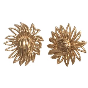 May include: A pair of gold-colored, sunflower-shaped clip-on earrings. Each earring features a detailed, openwork design with multiple petals radiating outwards from a central, textured core. The earrings are presented against a white background.