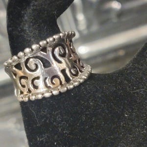 May include: Silver-toned ring with an openwork design featuring swirling patterns. The ring has a beaded border along the top and bottom edges. The ring is displayed on a black surface.