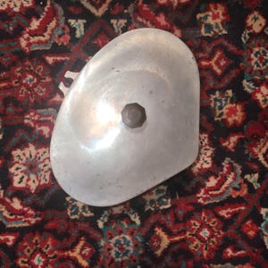 May include: A vintage, aluminum-colored metal object with a rounded, asymmetrical shape. It features a central, dark-colored, hexagonal knob. The object's surface reflects light, suggesting a polished finish. The background is a patterned rug with red, blue, and cream colors.