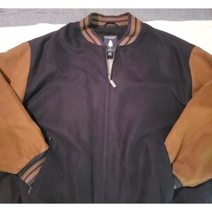 Pode incluir: Uma jaqueta bomber azul marinho com mangas de couro marrom. A jaqueta tem fecho de zíper completo e gola e punhos listrados. O nome da marca é visível na gola.