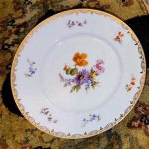 May include: White porcelain plate with a gold rim and floral design. The plate features a central bouquet of orange, purple, pink, and yellow flowers, with smaller floral accents around the edge. A decorative piece for serving.