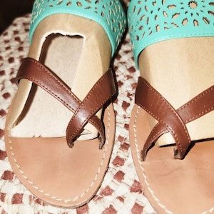 Op de afbeelding: Sandalen met bruine leren bandjes en lichtbruine zolen. De sandalen hebben een turquoise band met een decoratief uitgesneden ontwerp. De zolen zijn gestikt met witte draad.