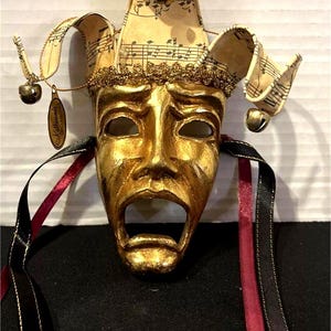 Puede incluir: Una máscara teatral dorada con la boca abierta y una expresión triste. La máscara presenta un gorro de bufón adornado con partituras y pequeñas campanas. Cintas negras y rojas cuelgan de la máscara, añadiendo un toque decorativo.