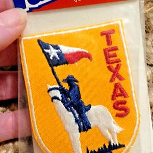 May include: Embroidered patch depicting the Texas state flag and a rider on a white horse. The patch has a yellow background with the word "TEXAS" in red. The top of the patch reads "Patches" and "Voyager Emblems".