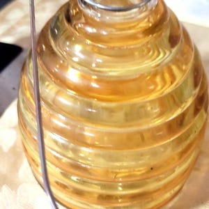 May include: A clear glass jar with a round, ribbed body and a clear glass stopper. The jar has a wire closure and is filled with a golden-yellow liquid. The jar sits on a cream-colored surface.