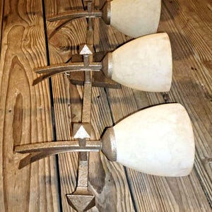 May include: A bronze wall sconce featuring four off-white, bell-shaped glass shades. The fixture has a vertical design with decorative metalwork. It is mounted on a wooden surface.