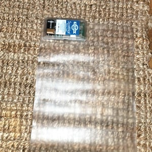May include: A clear, rectangular ScatMat with a small, clear plastic box containing a battery pack. The ScatMat is placed on a textured, tan rug. The box has the text "ScatMat" and "PetSafe".