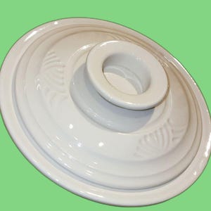 Könnte beinhalten: Weißer Keramikdeckel mit einem erhöhten Zentrum und einem dekorativen Design am Rand.