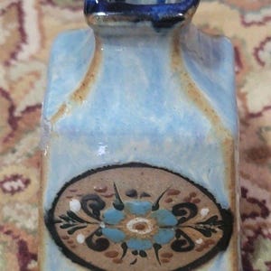May include: A small, rectangular ceramic vase with a light blue glaze and a dark blue rim. The vase features a brown and black floral design with blue and white accents. The vase is approximately 4 inches tall.