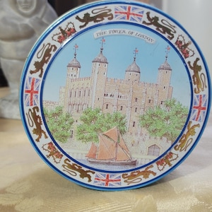 May include: A round tin featuring an illustration of the Tower of London, a boat, and the text "THE TOWER OF LONDON." The tin has a blue border with lions, flags, and a blue rim.