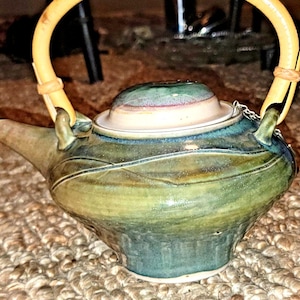 May include: A ceramic teapot with a bamboo handle. The teapot is glazed in shades of green and blue, with a cream-colored lid. The handle is a curved, light yellow bamboo. The teapot has a spout and a decorative design on the body.
