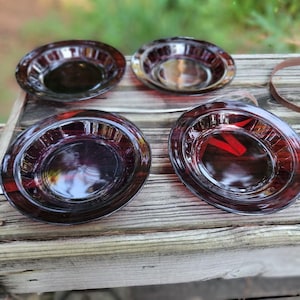 Può includere: Quattro piatti di vetro rosso scuro con un design circolare sono disposti su una superficie di legno stagionato. I piatti hanno un colore intenso e ricco e una qualità riflettente, suggerendo che siano fatti di vetro. La disposizione suggerisce un set di piatti da portata.