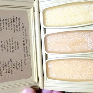 Może przedstawiać: Kompakt vintage Coty Sweet Earth Flower Fragrances. Otwarty kompakt ukazuje trzy prostokątne paski perfum w odcieniach beżu i brzoskwini. Tekst po lewej stronie opisuje zapachy: Hiacynt, Wiciokrzew i Ylang Ylang.