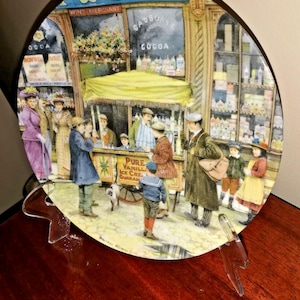 May include: Decorative plate featuring a vintage street scene with a shop labeled "& SON. WINE MERCHANT" and a yellow awning. The scene includes figures in period clothing, a vendor selling ice cream, and a dog. The plate is displayed on a clear stand.