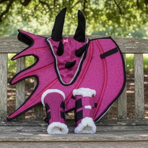 May include: A pink glittery dragon costume set, including wings, a headpiece with horns, and matching leg warmers with faux fur trim. The costume is designed for a child and is displayed on a wooden bench.