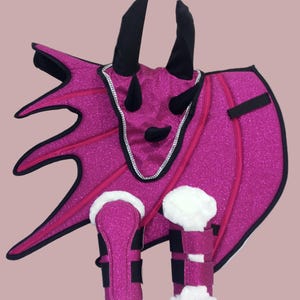 May include: A bright pink dragon costume set with glittery fabric, black horns, and white faux fur trim. The set includes a wing piece, headpiece, and leg warmers, perfect for a costume party or imaginative play.