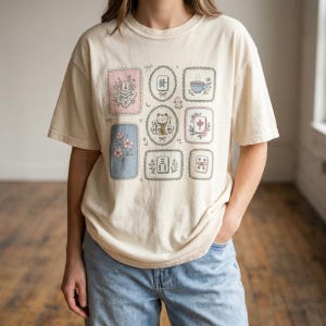 Puede incluir: Camiseta color crema con nueve ilustraciones enmarcadas, incluyendo una taza de té, flores y un gato de la suerte. La camiseta se combina con unos vaqueros azules.
