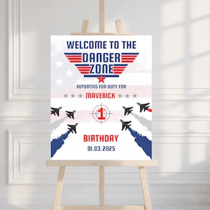 Könnte beinhalten: Ein weißes Schild auf einer hölzernen Staffelei mit dem Text "WELCOME TO THE DANGER ZONE" in Blau und Rot. Das Schild enthält eine Grafik eines Jets und die Worte "REPORTING FOR DUTY FOR MAVERICK BIRTHDAY 01.03.2025". Der Hintergrund zeigt eine verblasste amerikanische Flagge.