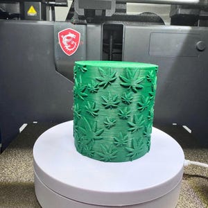May include: A green, cylindrical object featuring a cannabis leaf design, possibly 3D printed. It sits on a white rotating platform, with a 3D printer visible. The leaves are raised and detailed.