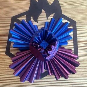 Puede incluir: Objeto decorativo impreso en 3D con un diseño en capas en tonos azules y morados. Se encuentra sobre una forma geométrica negra y una superficie de madera. El diseño presenta un patrón radial.
