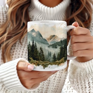 May include: White ceramic mug with a watercolor design of a mountain range with fog and trees. The mug has a handle on the right side.