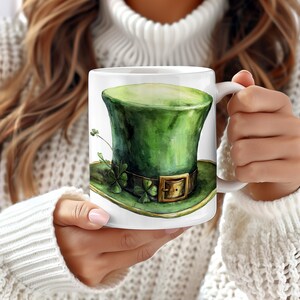 May include: White ceramic mug with a green leprechaun hat with a gold buckle and shamrocks on the brim. The hat is painted in a watercolor style.