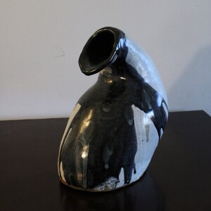 Puede incluir: Un jarrón de cerámica blanco y negro con una forma abstracta única. El jarrón tiene una abertura ancha en la parte superior y una base estrecha. El esmalte es desigual, creando un efecto moteado.