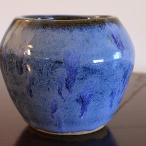 Può includere: Un piccolo vaso in ceramica blu con un bordo marrone e una glassa screziata. Il vaso ha una forma arrotondata e un labbro leggermente svasato.