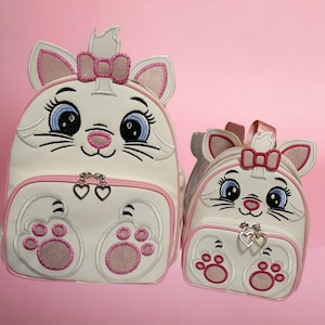 Könnte beinhalten: Zwei weiße katzenförmige Rucksäcke mit rosa Akzenten. Der größere Rucksack hat eine rosa Schleife oben und der kleinere Rucksack hat eine rosa Schleife oben auf dem Kopf. Beide Rucksäcke haben gestickte Augen, eine Nase und Schnurrhaare.
