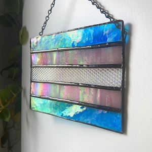 May include: Stained glass panel in the shape of the transgender pride flag. The panel features blue, pink, and white iridescent glass, framed with black metal and suspended by a chain.