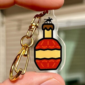 May include: A clear acrylic keychain featuring a cartoon illustration of a bourbon bottle with a gold-colored clasp. The bottle design includes red, yellow, and brown colors, with a small silhouette of a horse and rider on top.