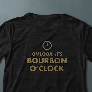 Puede incluir: Camiseta negra con un reloj dorado y el texto "OH LOOK, IT'S BOURBON O'CLOCK"