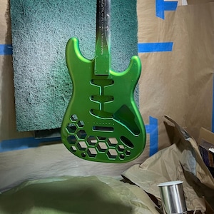 May include: A green electric guitar body with a honeycomb design, suspended in a workshop. The guitar is painted in a vibrant green hue, with a metallic sheen. The background includes a green textured surface and painter's tape.