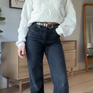 May include: A woman wearing a white cable knit jumper with a high neck and black jeans. She is wearing a silver belt with a buckle.
